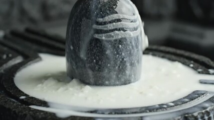 Close up of a sacred dark stone lingam being bathed in creamy white milk with a stone pedestal and soft focus background in a sacred space