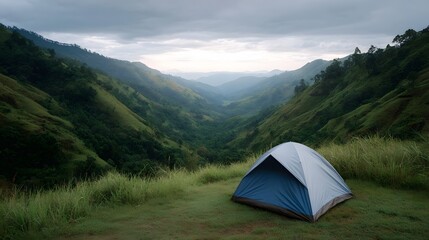 A camping tent is set on a grassy hill overlooking a lush mountain valley under an overcast sky