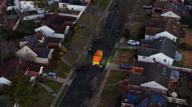 An aerial view of a residential neighborhood during a cloudy morning, following a yellow sanitation truck and workers picking up the trash. The camera is tilted down and dolly in.