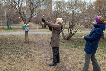 Elderly woman learning archery in autumn park with younger woman. Intergenerational family leisure and active healthy pastime. Outdoor exercise for senior.