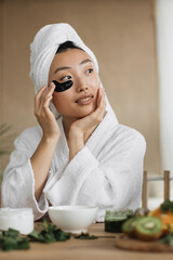A young Asian woman in a white bathrobe and towel applies black under-eye patches for skincare
