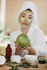 Fototapeta premium A young Asian woman with a white towel wrapped around her head and a green clay mask on her face holds a green pomelo fruit while surrounded by natural beauty products