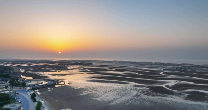 Aerial time-lapse of tidal flats at sunrise, winding water channels and golden reflections under a soft morning sky, Rizhao City, Shandong Province, China.