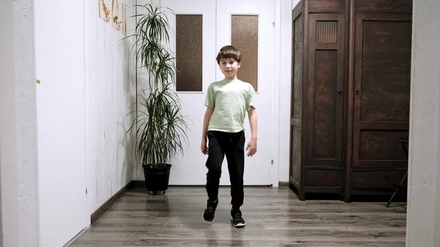 Caucasian 9 year old boy standing and alternately raises legs up. Morning exercises of a schoolboy. Warm-up for the whole body at home. Healthy children lifestyle. Good mood. Body care. 4K footage.