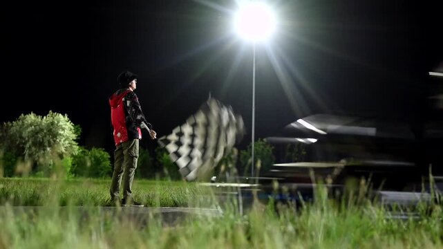 Night race marshal waving checkered flag under bright floodlight, blurred kart streaks past finish on wet