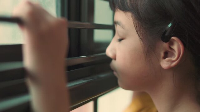 Close up side profile of sad Asian girl looking out window with selective focus. Lonely child face details with blurred hand holding bars in foreground, concept of waiting, depression, or boredom.