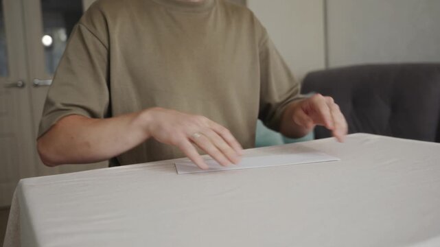 Medium static shot from table level of an adult man aligning and straightening the paper airplane base. Camera focuses on hands and paper, calm home interior in the background. High quality 4k footage