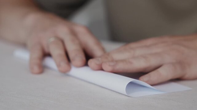 Close-up side view at table height showing an adult man smoothing a folded sheet of paper with both hands while preparing a paper airplane. Focus on flat surface and crease alignment. High quality 4k