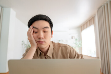 Young Asian man tired bored sad upset fatigued working on laptop in living room at home