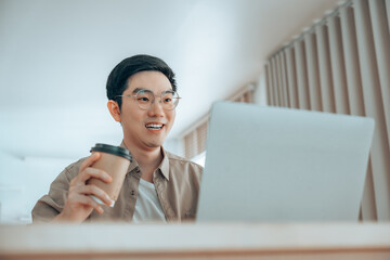 Happy young Asian man working on laptop in living room. Freelance worker male sitting at desk writing notes while watching webinar, studying online