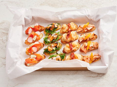 Different baked bread slices are arranged on a tray, each topped with various ingredients such as seafood, vegetables, and spreads. The setting shows a food gathering