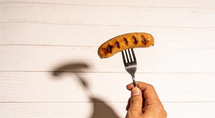 Man holding fork with grilled sausage on white background, closeup. Barbecue food