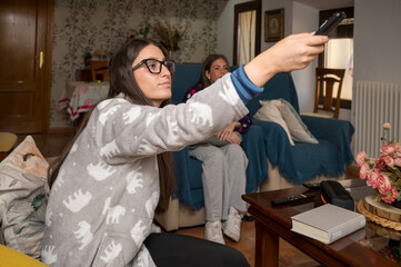 Teenage girl pointing remote control towards the TV while sitting with her friend in the living room during a cozy afternoon at home