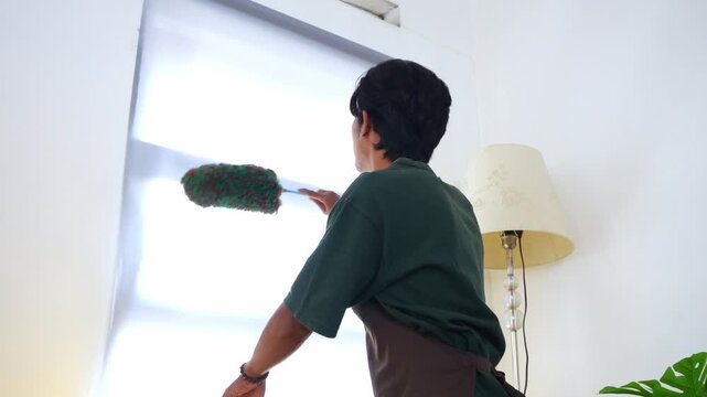 Young asian Man uses feather duster to clean white roller blinds in home