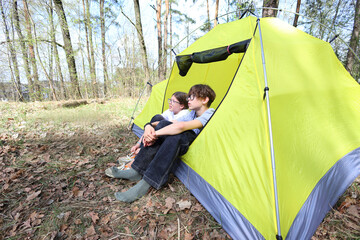 Two kids sit cozily at the entrance of their bright yellow tent, gazing into the forest. A quiet moment of shared wonder and companionship on a sunny spring camping trip.