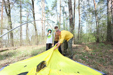 A father and two kids set up a bright yellow tent in a sun-dappled forest. A moment of teamwork, learning, and family bonding on an outdoor adventure.