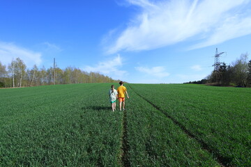 Two children walk hand-in-hand through a vibrant green field under a bright blue sky. A peaceful, hopeful scene of childhood exploration and connection with nature.