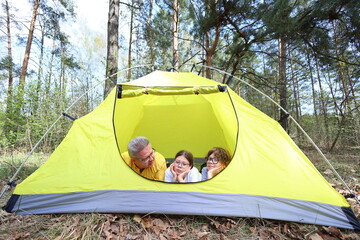 A father and two kids rest inside their yellow tent, gazing out at the forest. A quiet, cozy moment of shared wonder and connection on a sunny spring camping trip.