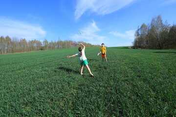 Two kids play tennis with plastic rackets in a wide green field under a sunny blue sky. Pure, active childhood joy and carefree outdoor fun.