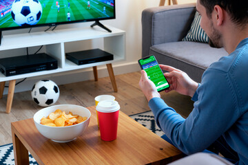 Caucasian young adult man sitting on sofa watching soccer match on television while holding smartphone with sports betting app, bowl of potato chips and drinks on wooden coffee table