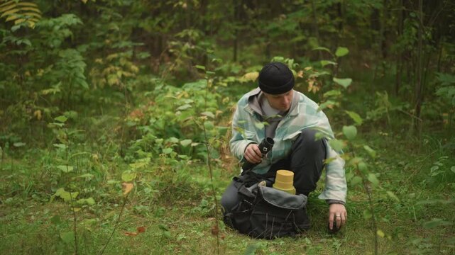 Hiker kneeling unpacks backpack pulling gear and inspecting items adjusts straps, selects rope and tool, practices campcraft techniques in dense green forest, focused preparation for shelter or trail