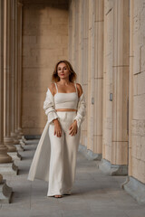 Fototapeta premium Woman White Outfit Columns Fashion - A woman wearing a white outfit stands in a hallway with columns. The photo was likely taken for a fashion magazine or blog.