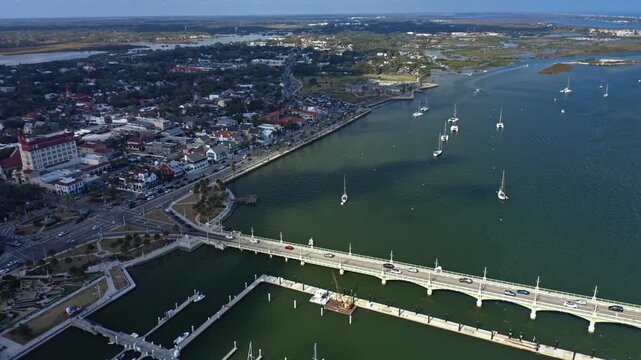 Coastal urban area with a bridge crossing water and sailboats anchored near docks, surrounded by city streets, buildings, and green spaces along the shoreline in a temperate setting.