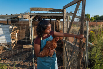 Young farmer girl closing chicken coop with hen in her arms