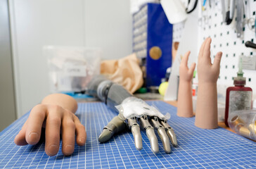Tools hang above the work surface used to create and customize prosthetics. Various artificial hands and a partial arm rest on a gridded workbench in a well lit workshop