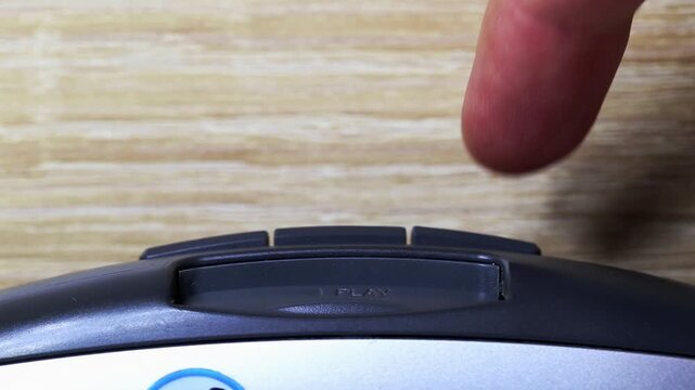 Detail view of index finger activating play button of gray and silver electronic device against wooden background. Action of starting or resuming audio or video playback.