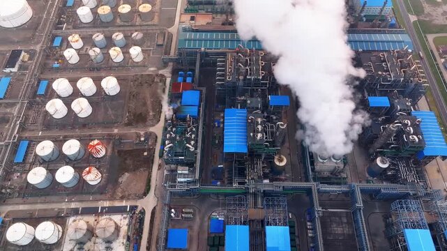 Aerial view of chemical plant with smokestack emitting white vapor and numerous storage tanks