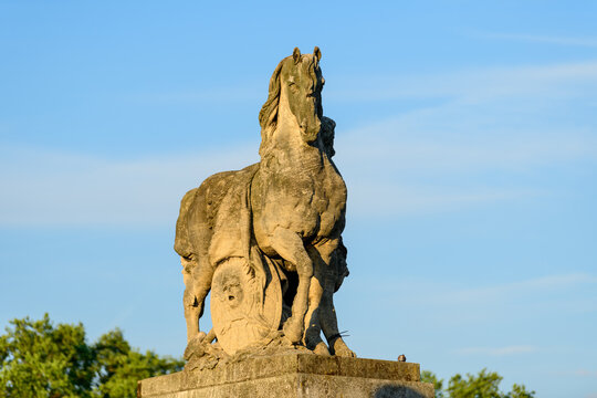 Impressive stone sculpture of a horse atop a pedestal on Pont de Iena in Paris, illuminated by warm sunlight against a clear blue sky and green foliage.