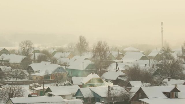 Winter morning view of rural village. Snow blankets roofs of houses and bare trees. Smoke from chimneys shows warmth on cold, frosty dawn. Hazy sky.