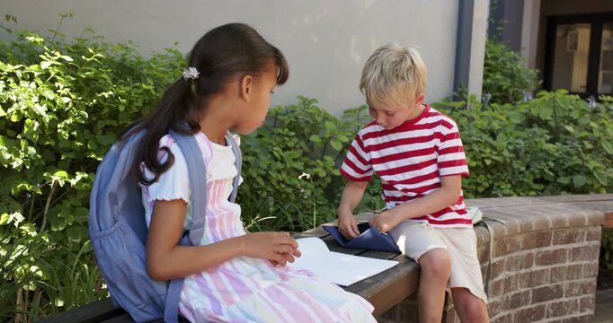 Two children sitting on wooden bench by brick wall, boy opening pencil case, writing homework