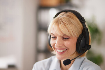 Woman sits at an office desk wearing a headset. She smiles as she talks, providing assistance to customers. The setting has shelves in the background filled with items.