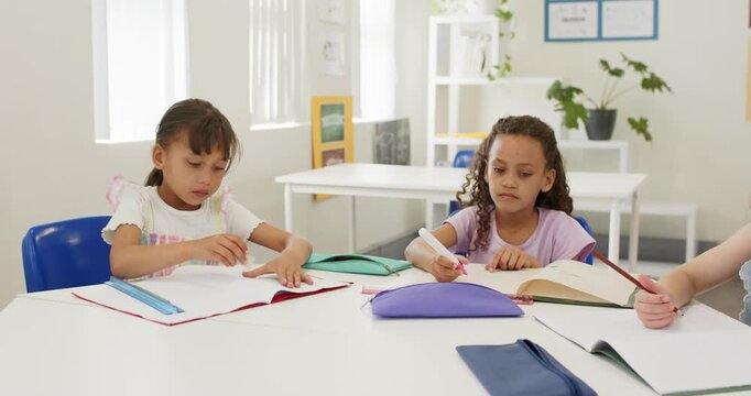 Diverse children at table left glancing at camera opening book middle marking right writing work