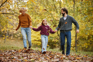 A family of three takes a joyful weekend stroll through a beautiful autumn park, playfully holding hands and laughing as they step on colorful leaves.