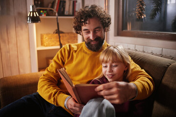 A parent and child sit on a comfortable sofa in a warmly lit room, enjoying a book together during...