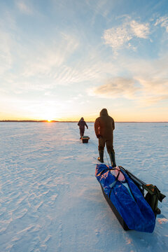 Two anglers trek across a frozen lake at sunrise, pulling sleds with an auger, rods, and a tarp shelter. Long shadows and warm low light mark deep winter morning.