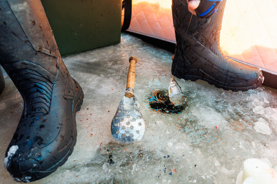 Ground level view shows gloved hand holding a panfish on a handline beside a skimmer, slushy drilled hole, wet boots, and quilted orange wall during a winter day.