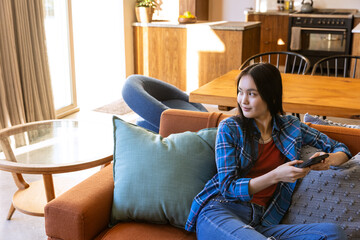 Obraz premium Asian woman sitting on orange sofa holding smartphone in sunlit living area