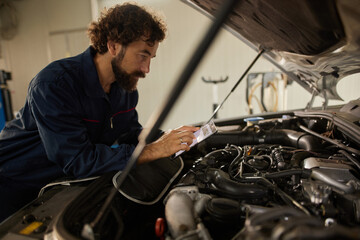 A car mechanic is focused on a service manual while examining the engine of a vehicle in a repair shop. Tools and equipment are visible in the background.