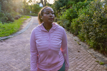 Fototapeta premium African american woman walking through park on brick-paved path wearing jacket, earbuds