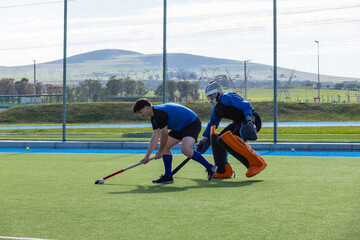 Male field hockey players controlling ball on turf with stick and helmeted padded goalkeeper