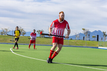 Diverse male field hockey players running on artificial turf with hockey sticks in uniforms