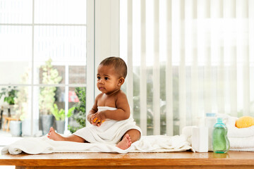 A baby wrapped in a towel sits on a changing table by a window, surrounded by baby care products in...