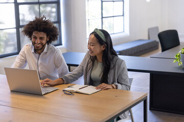 Diverse coworkers working at office desk with laptop, notebook, pen, eyeglasses, potted plant