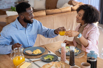 Diverse couple toasting glasses of orange juice while sharing breakfast at dining table