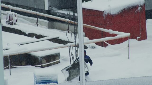 Worker in blue winter suit pushing large heavy object through deep snow on outdoor industrial site. Winter storm with falling snow creates challenging conditions for manual labor.