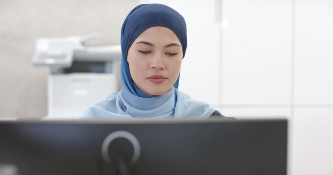 African American woman wearing hijab reviewing monitor at office, glancing to printer to check job